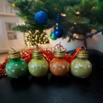 holiday product photo of a white gift box open with tinsel and scattered confetti. inside are four christmas ornaments filled with kratom powder.